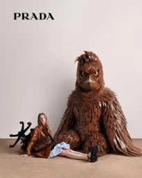 Woman in brown leather jacket seated next to a large brown sculptural bird figure against a light background