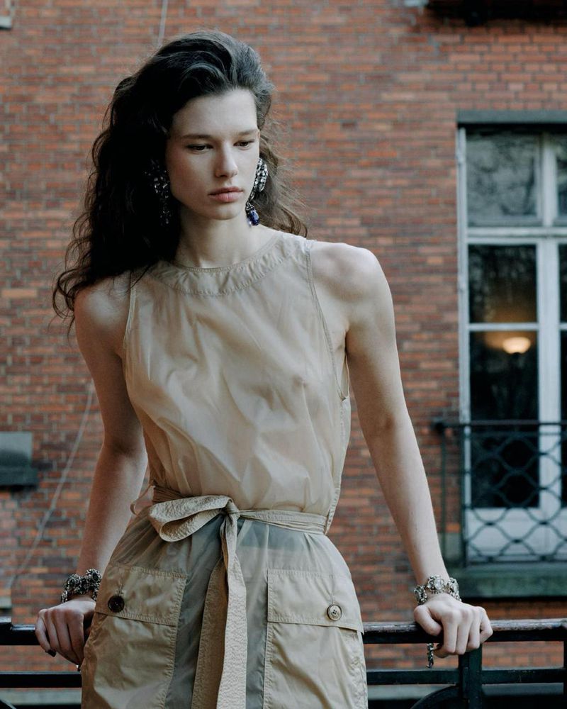 Brunette woman in sheer beige top and skirt with a tie, posing on a balcony with a brick building in the background, wearing jewelry.