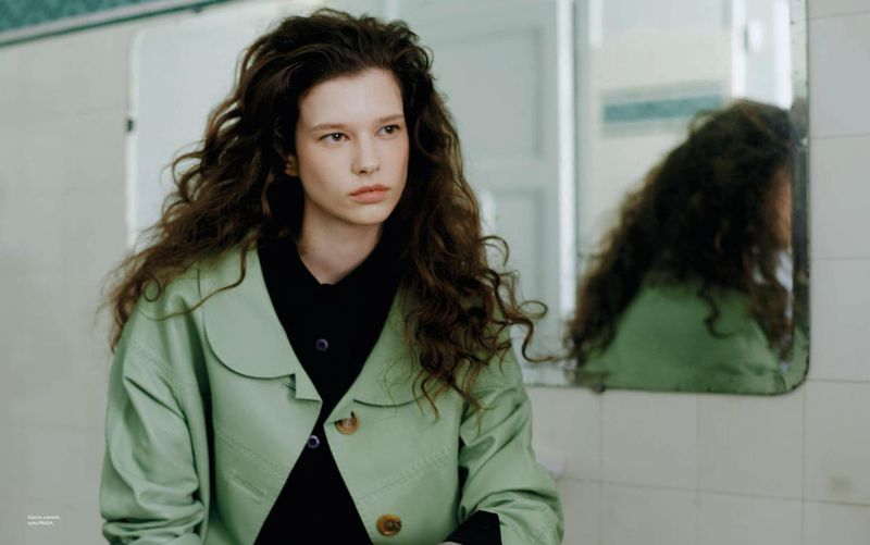 Woman with curly brown hair wearing a green jacket in front of a mirror in an indoor setting, medium shot, eye-level angle.