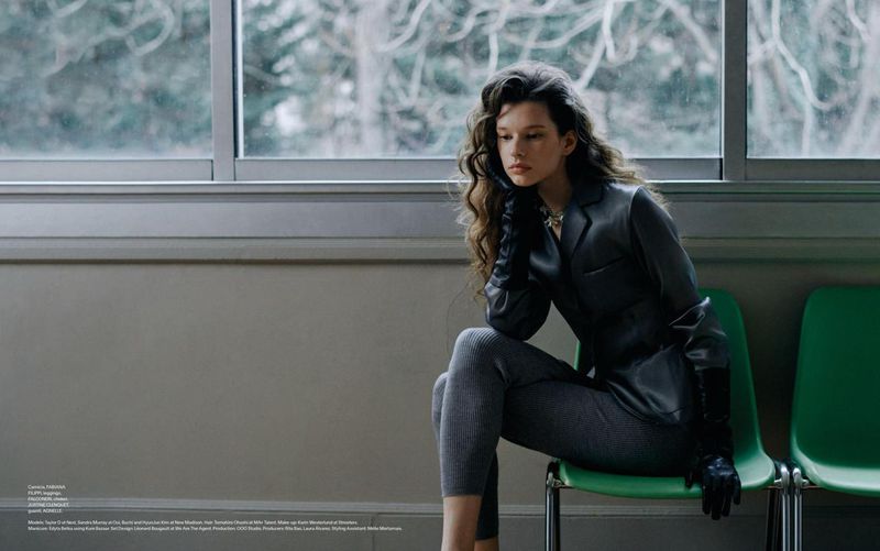 Woman with long curly hair poses while sitting on a green chair, wearing a grey suit with long black gloves and silver necklace near a window.