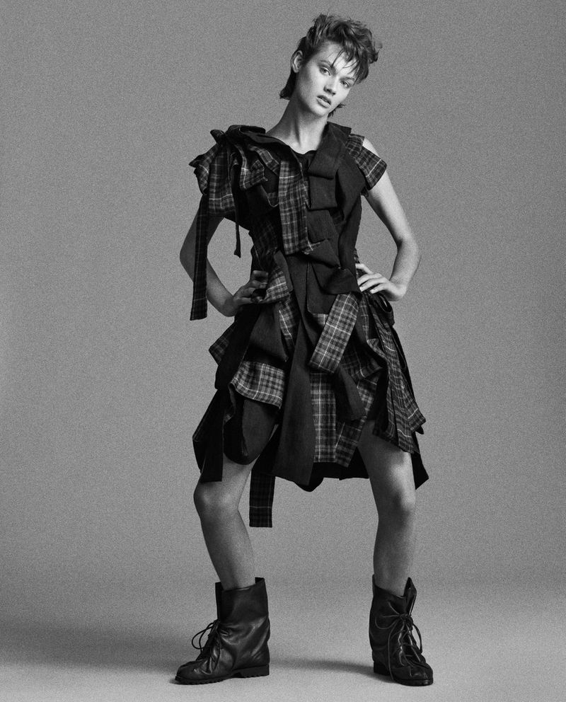 Monochrome fashion shot of a young adult woman in a deconstructed plaid dress and platform boots, hands on hips in a photo studio.