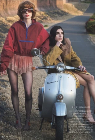 Two women in stylish outfits pose with a silver Vespa scooter on a paved road with grassy and building backgrounds, creating a vintage fashion editorial.
