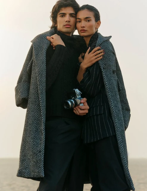 Two fashion models pose outdoors in overcoats. A man holds a vintage camera with a flash attachment. Neutral background with soft lighting.
