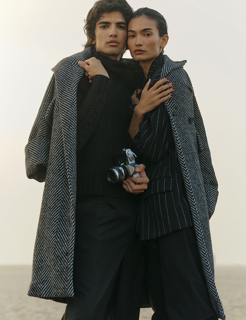Stylish couple in outerwear poses outdoors. The man holds a vintage camera. Toned lighting enhances fashion appeal with a neutral background.