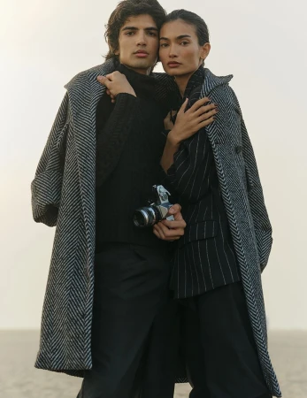 Stylish couple in outerwear poses outdoors. The man holds a vintage camera. Toned lighting enhances fashion appeal with a neutral background.