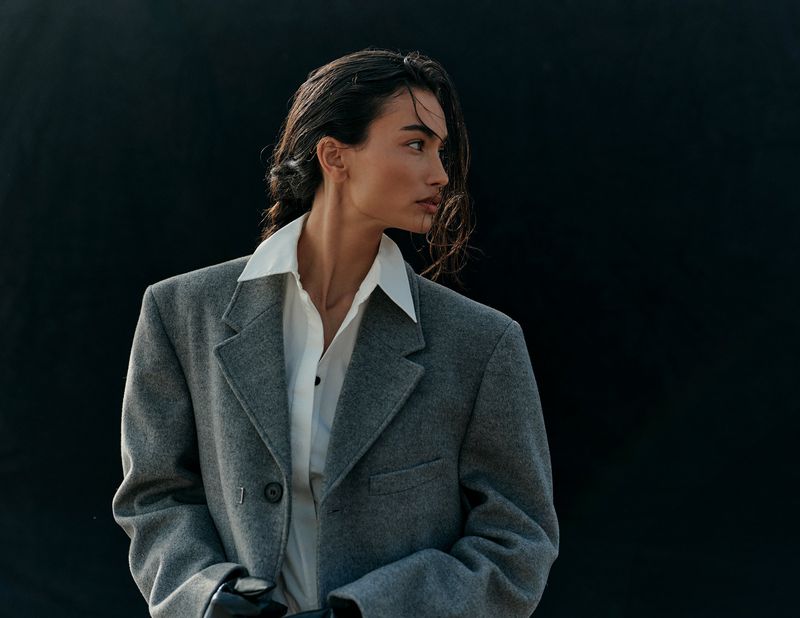 Elegant woman in oversized grey blazer and white collared shirt poses against a dark background in a fashion-forward studio portrait.