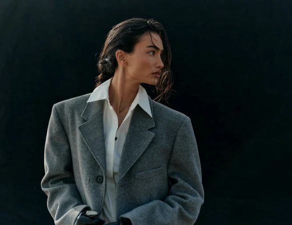 Elegant woman in oversized grey blazer and white collared shirt poses against a dark background in a fashion-forward studio portrait.