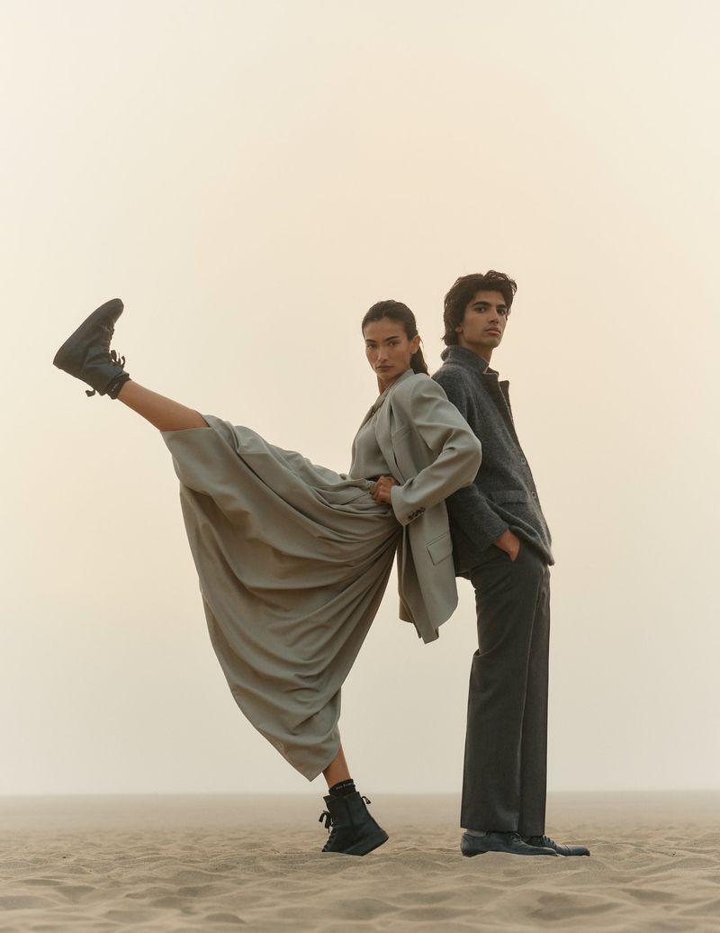 Two models pose on a sand dune. A woman in a matching jacket and skirt kicks one leg in the air, while a man stands behind her.