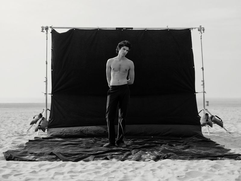 Black-and-white portrait of a shirtless male model standing in front of a large black backdrop on a sandy beach during a photoshoot.