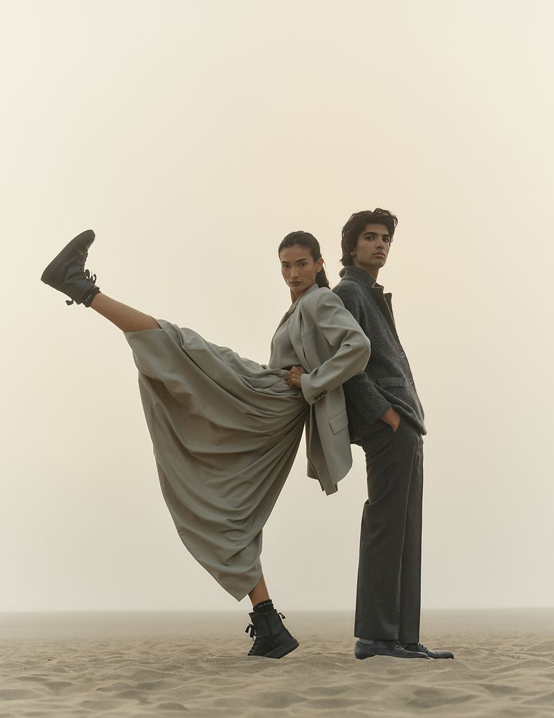 Two fashion models strike a pose back-to-back on a sandy surface under a hazy sky. One model is lifting their leg wearing a boot.
