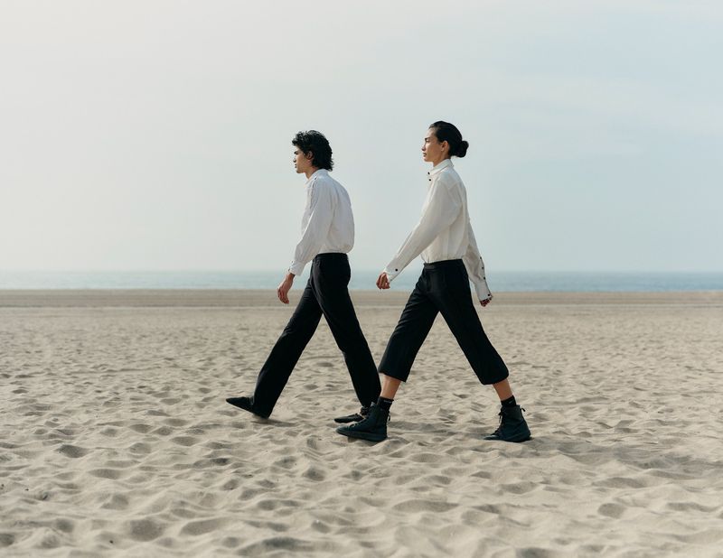 Two people in white shirts and dark pants walk on a sandy beach with a distant ocean view under a bright, overcast sky.