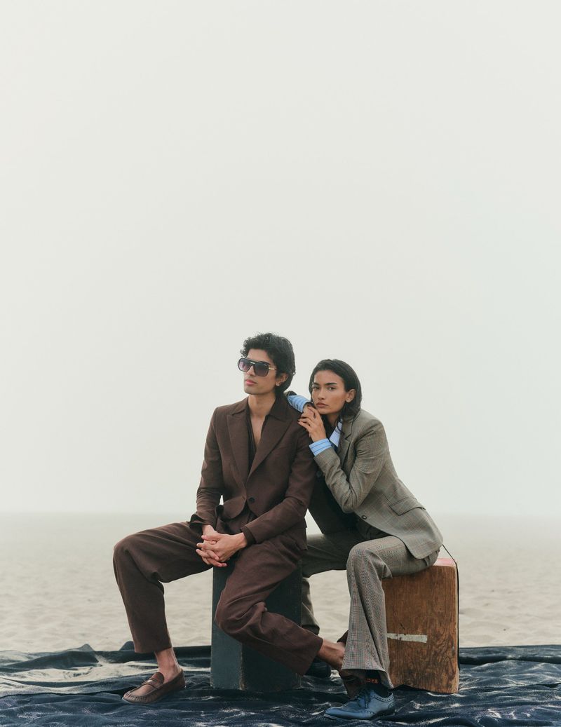 Two fashion models in brown suits pose on a box against a muted sky and landscape for a fashion editorial in a studio.