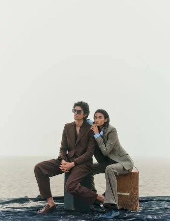 Two fashion models in brown suits pose on a box against a muted sky and landscape for a fashion editorial in a studio.
