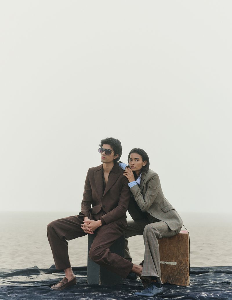 Two fashion models in suits pose on a wooden box in a sandy outdoor setting. One model wears sunglasses and brown loafers.