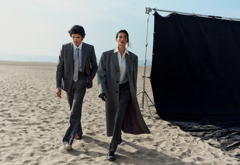 A fashionable couple in grey suits walks on a beach during a photoshoot with a black backdrop under a bright, clear sky.