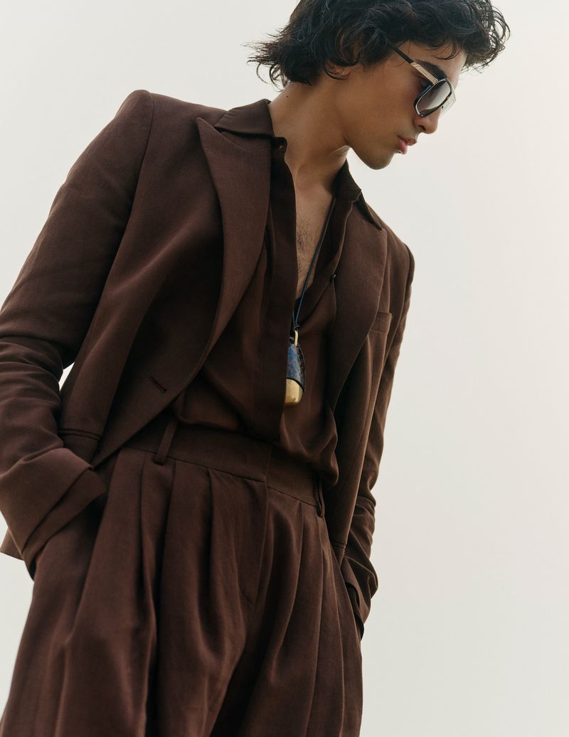 Man in brown linen suit and sunglasses poses against a white backdrop. Shirt is open, revealing a necklace.