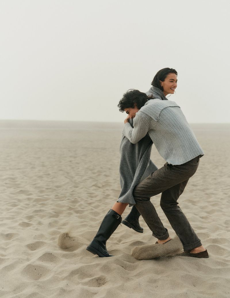 Two people in sweaters playfully interacting on a sandy beach with an overcast sky, creating a candid and relaxed scene.