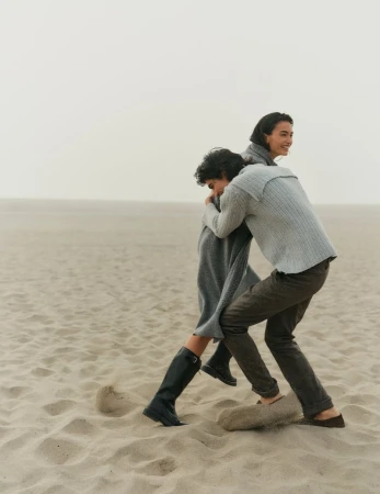 Two people in sweaters playfully interacting on a sandy beach with an overcast sky, creating a candid and relaxed scene.