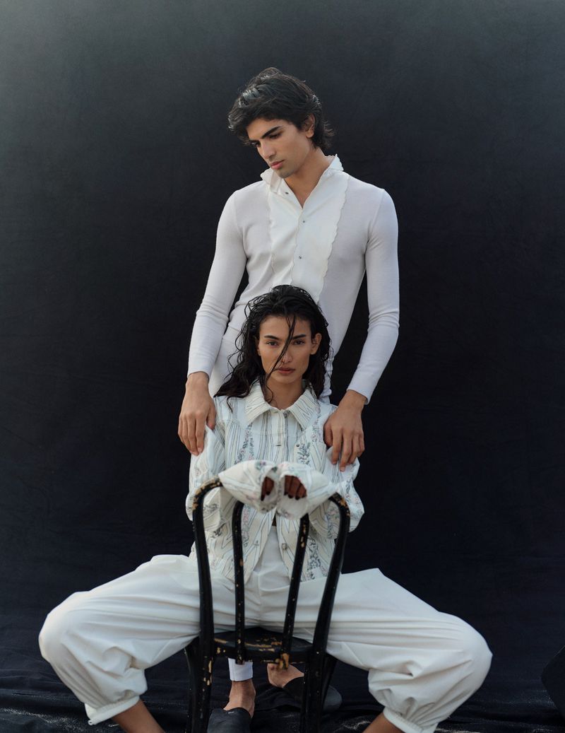 Two fashion models in studio posing. Man stands behind the other who is sitting on a black chair. They wear white tailored outfits.