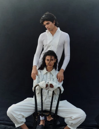 Two fashion models in studio posing. Man stands behind the other who is sitting on a black chair. They wear white tailored outfits.