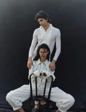 Two models pose in a studio setting. One sits in a black chair wearing white pants and a white, collared shirt with floral embroidery. The other stands behind.