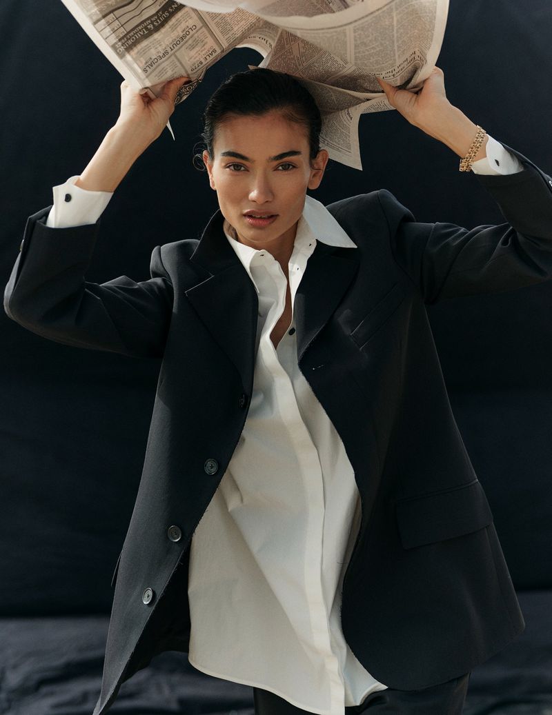 An adult woman in a black blazer and white button-down shirt holds a newspaper over her head against a dark background, indoors.