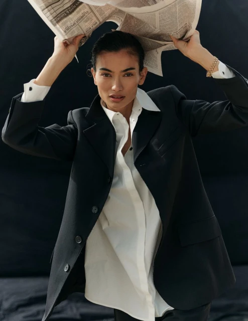 An adult woman in a black blazer and white button-down shirt holds a newspaper over her head against a dark background, indoors.