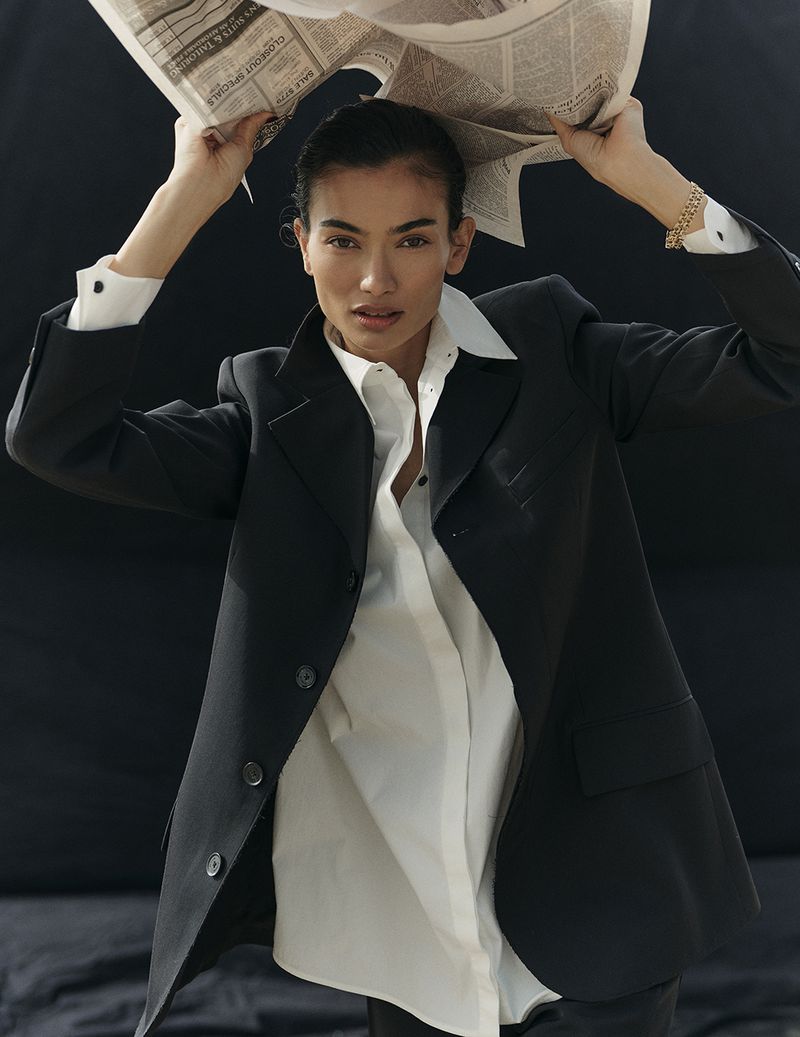 Adult woman in black blazer and white shirt poses with newspaper above head in a studio with dark background and soft lighting.