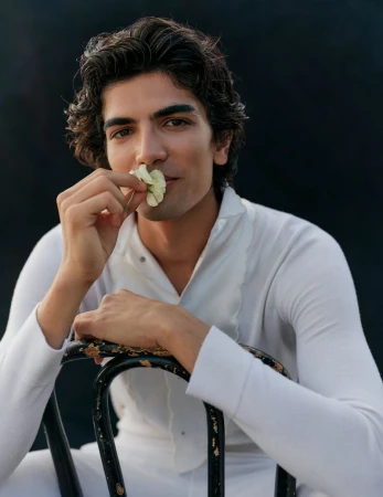 Man in white shirt holding a small flower to his nose while sitting on a chair against a dark background