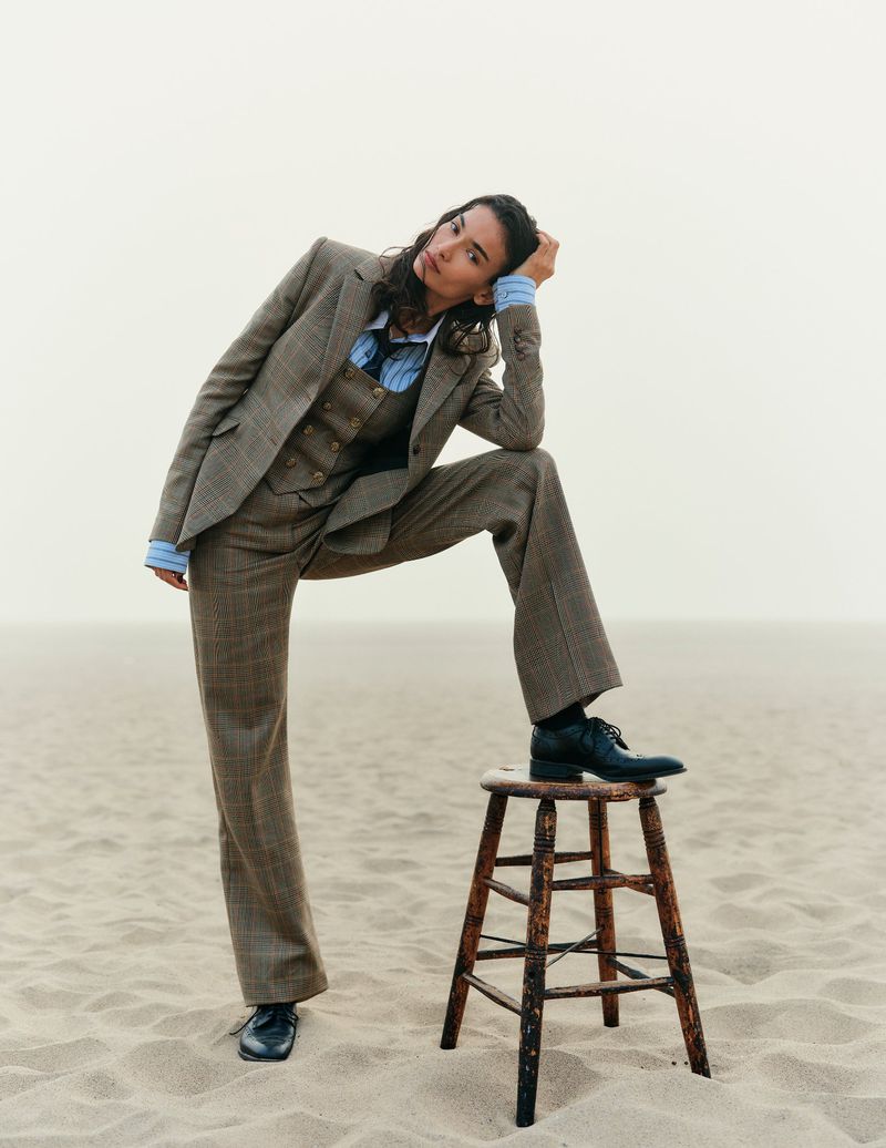 Fashion shot of a woman in a brown suit posing with one foot on a wooden stool in a desert landscape with a diffused, overcast sky.