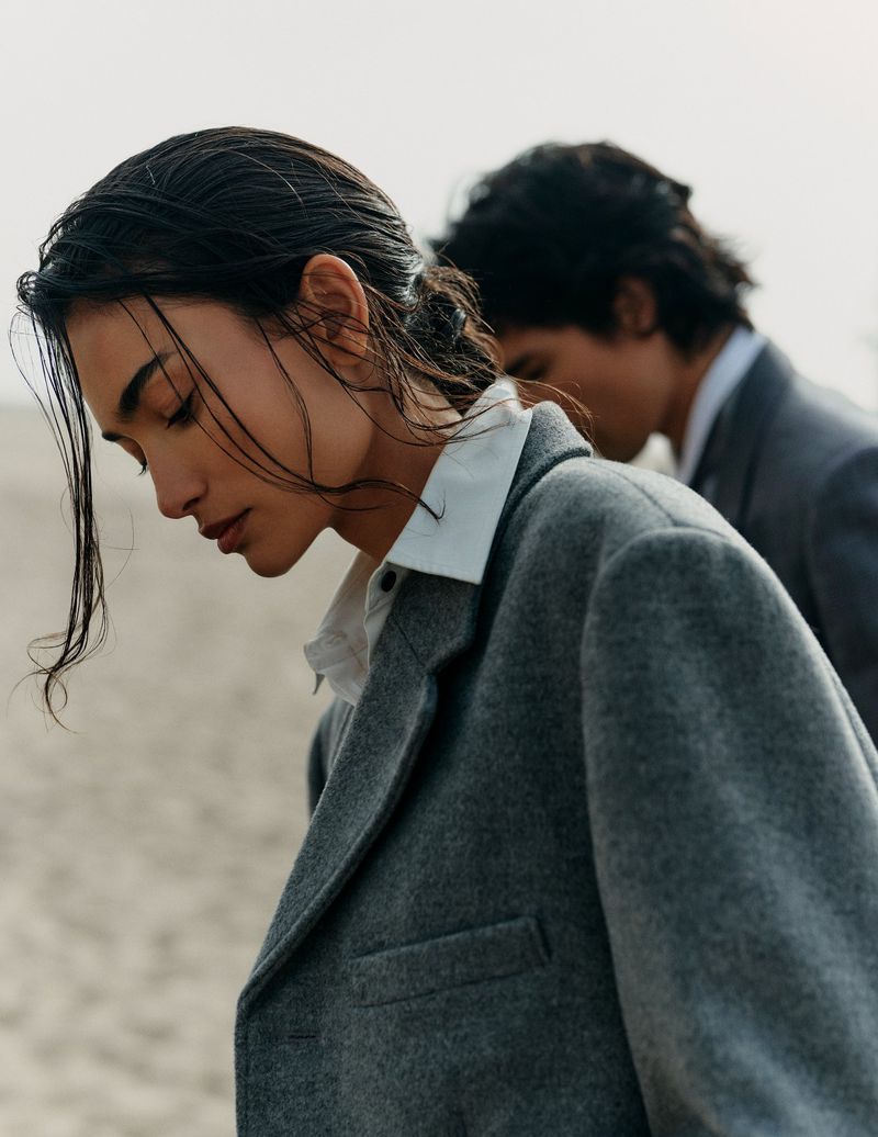 A woman and man in a close-up fashion shot are seen on the beach. The woman in the foreground is wearing a grey overcoat with wet hair.