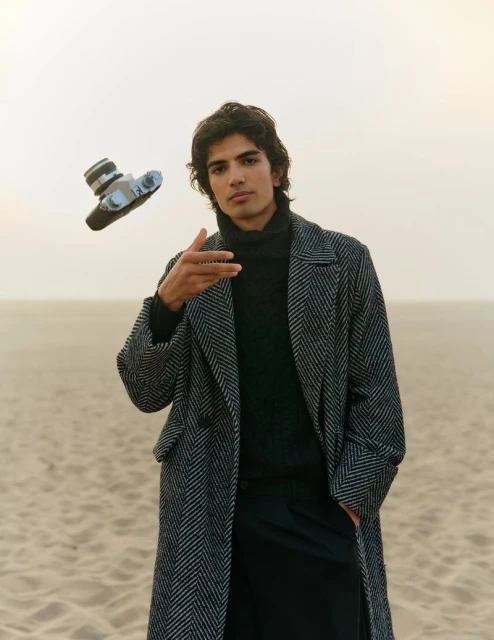 Stylish young adult male poses outdoors in a herringbone coat with a vintage camera levitating near him against a sandy beach backdrop.