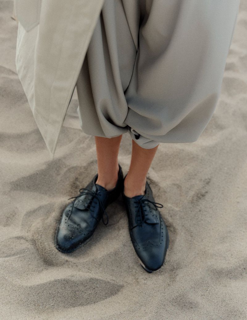 Person in baggy gray pants and black dress shoes standing on sand. Neutral and minimalist fashion on a beach.