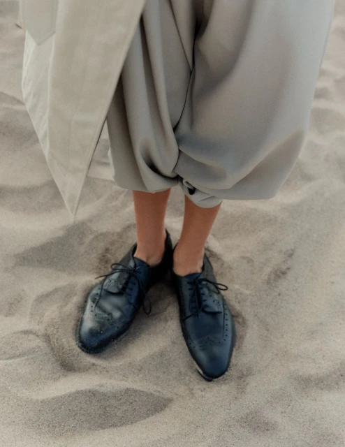 Person in baggy gray pants and black dress shoes standing on sand. Neutral and minimalist fashion on a beach.