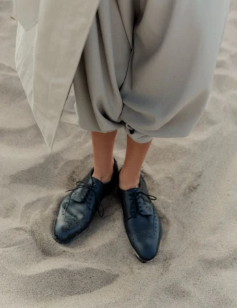 Person in baggy gray pants and black dress shoes standing on sand. Neutral and minimalist fashion on a beach.