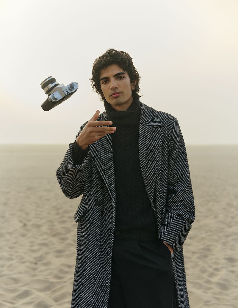 Young man in herringbone coat throws vintage camera on sandy beach with soft light, dark turtleneck, pants, and overcast sky.