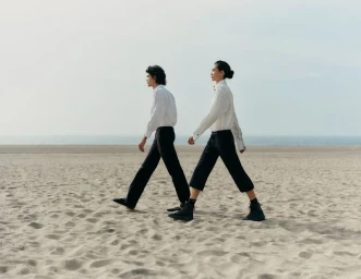 Two models wearing white shirts and dark trousers walking together along a sandy beach under an overcast sky.