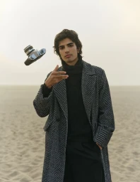 Man wearing a herringbone coat over a black turtleneck on a sandy beach while a vintage camera floats in mid-air