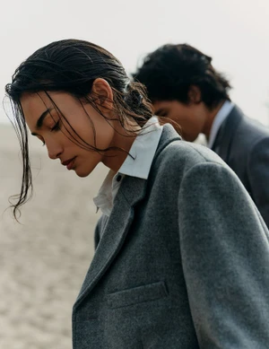 Model with damp hair wearing a grey wool blazer and white shirt outdoors with another person in the background