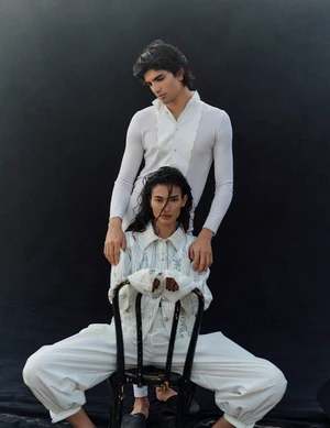 Two models posing in white outfits against a dark background, with a seated woman in front and standing man behind