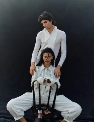 Two models wearing white outfits posed together against a dark background, with a woman seated on a black chair.