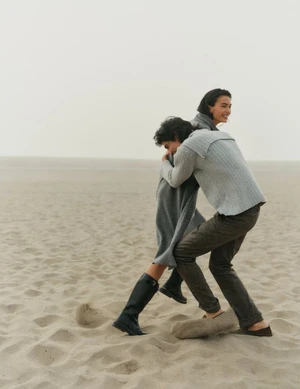 Two people embracing on a sandy beach under an overcast sky, captured in a candid moment for Vogue Scandinavia.