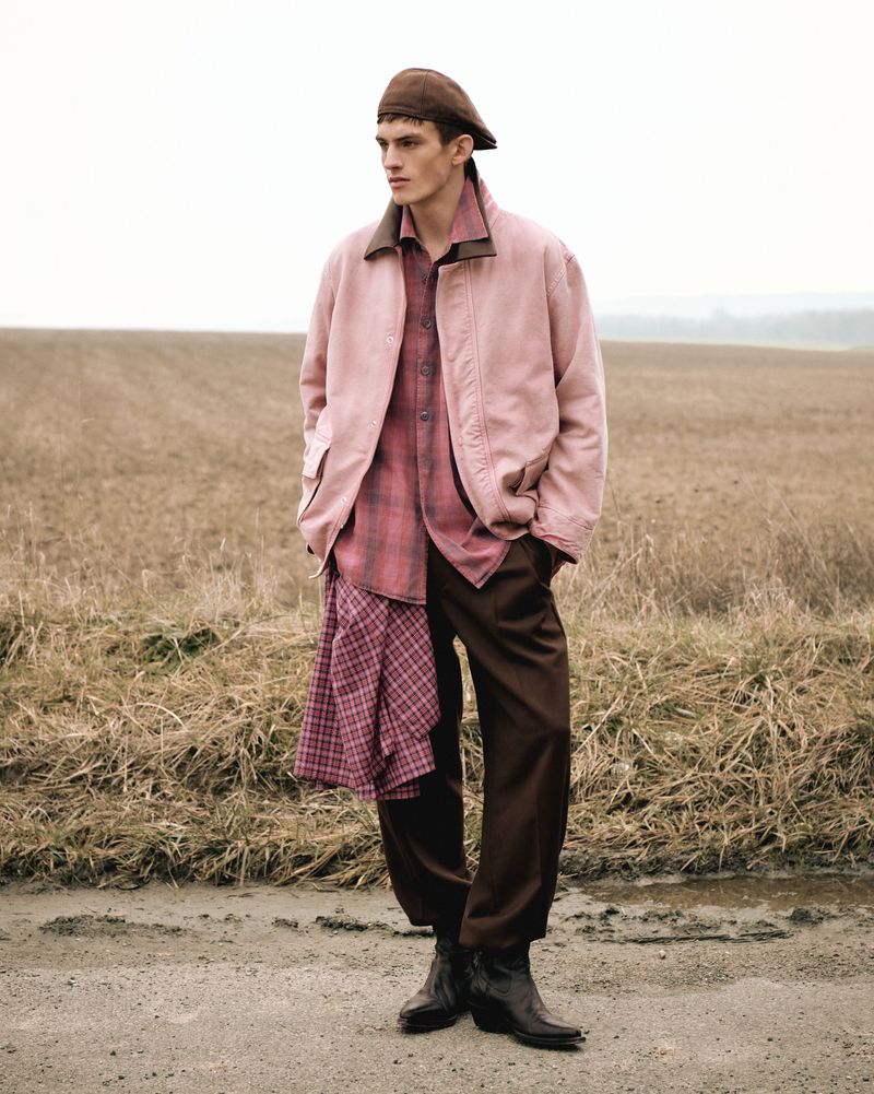 Male fashion model poses in a rural setting wearing a pink jacket, plaid shirt, brown pants and boots, and a brown cap outdoors.