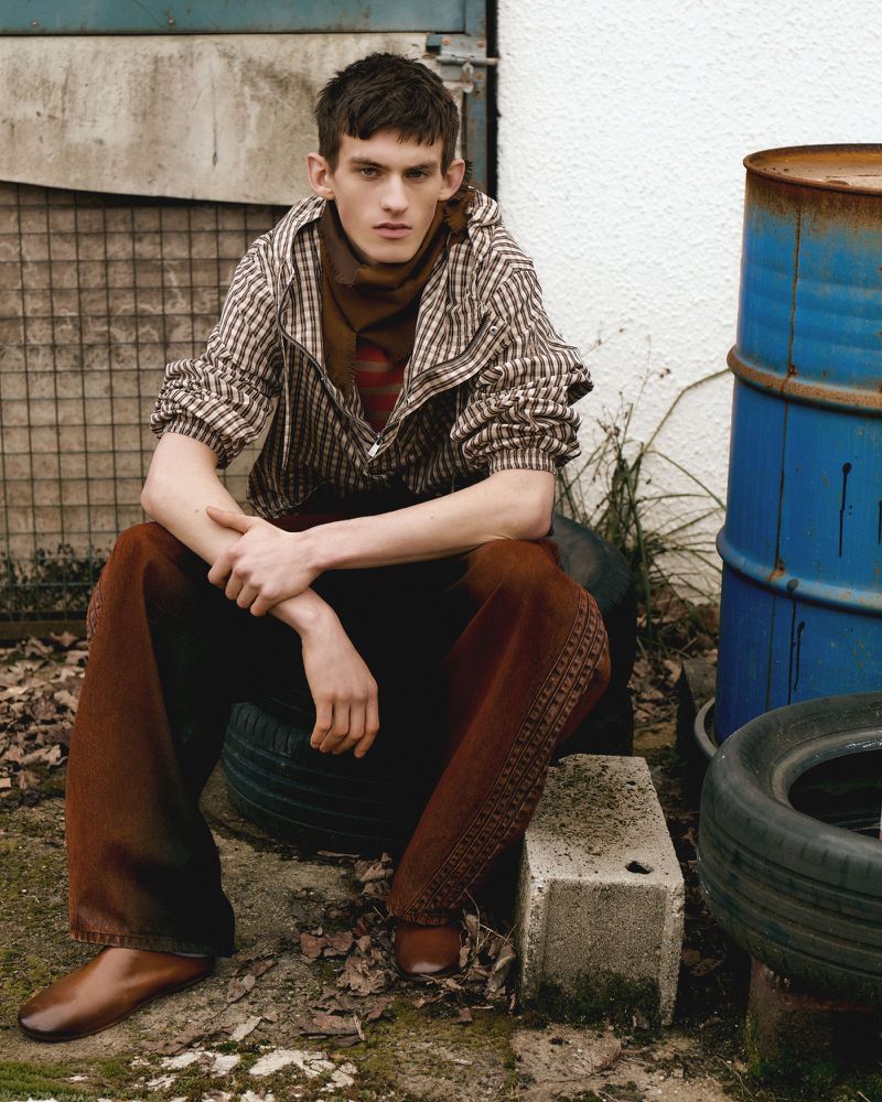 Young man in a striped jacket sits casually among tires and debris, creating an edgy and slightly gritty outdoor fashion portrait.