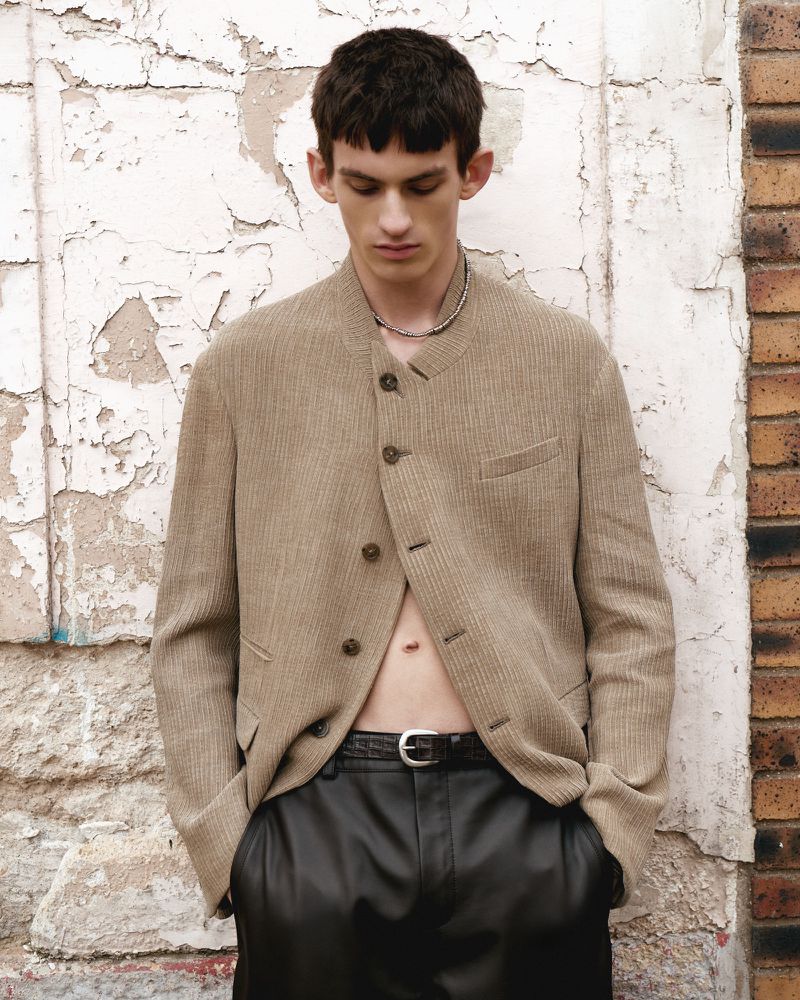 Male fashion model poses in tan jacket and black pants against textured wall for an editorial fashion shot.