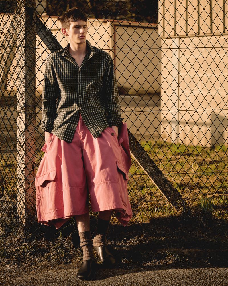 Young man in a checkered shirt and pink shorts stands outdoors against a chainlink fence, looking off to the side in a fashion shot.
