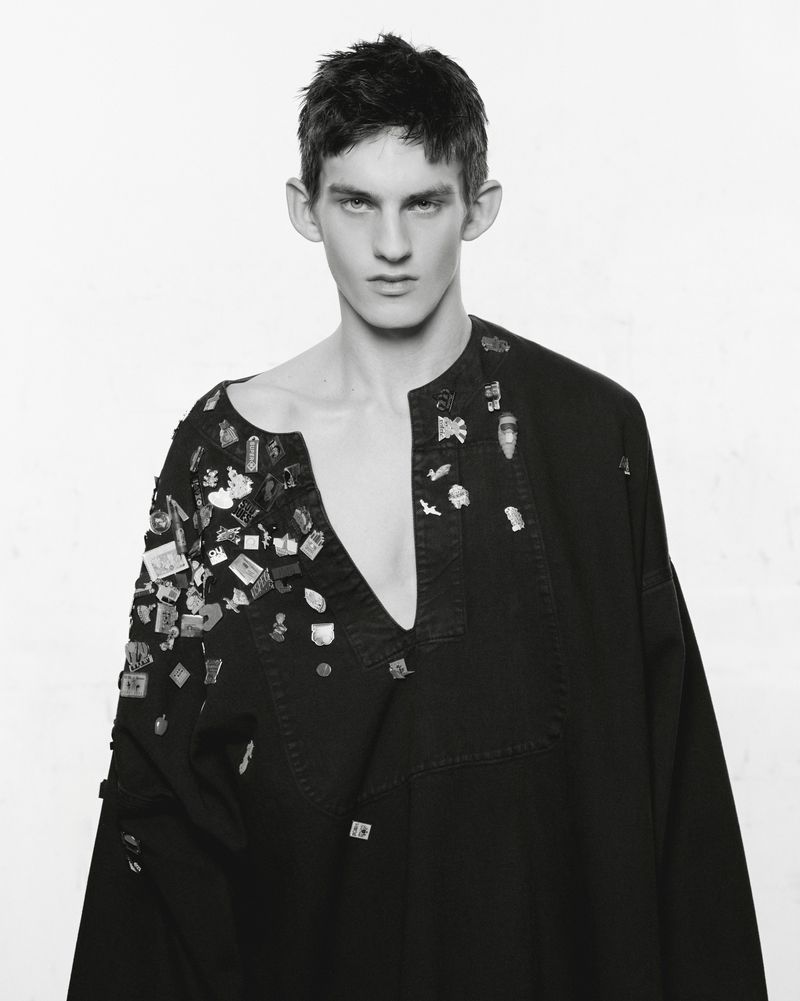 Studio portrait of a young adult man in a black, wide-necked shirt adorned with pins, against a bright white background in black and white.