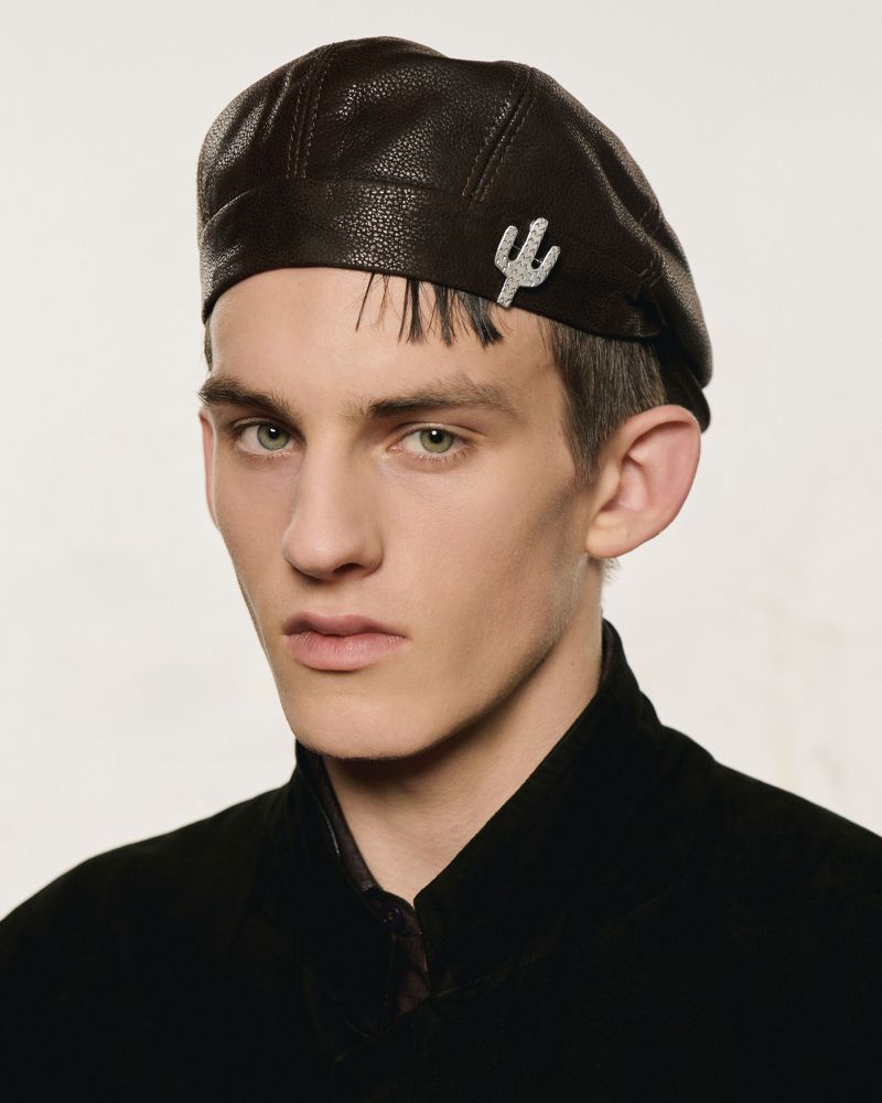 Portrait of a young adult man wearing a brown leather beret with a decorative cactus pin, styled in a black collared shirt.