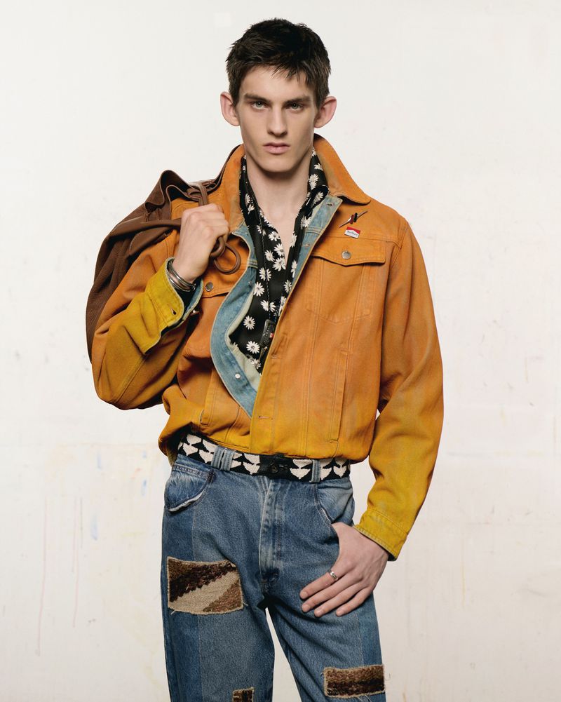 Male fashion model poses in denim jeans, orange jacket, and floral scarf in a studio setting with a duffel bag over his shoulder.