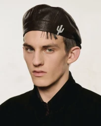 Close-up portrait of a male model wearing a dark leather beret with a crystal cactus brooch and a black collared top against a plain wall.